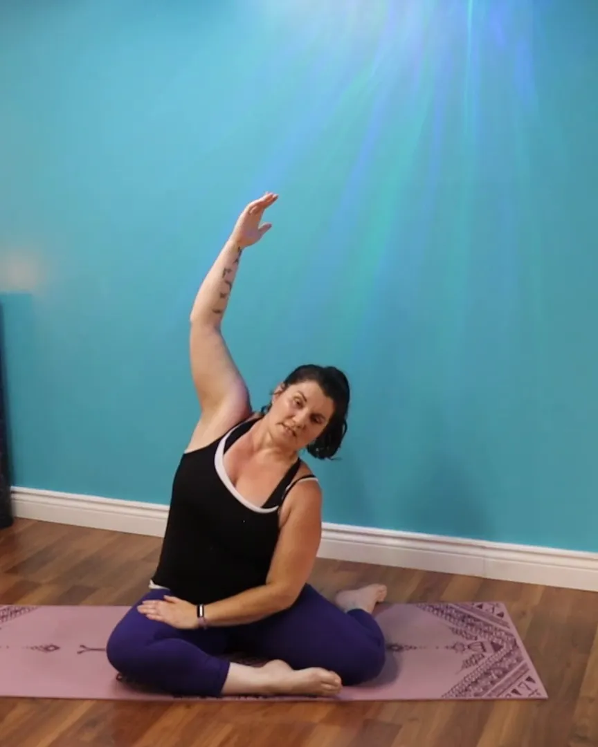 Angélique seated on her mat in a side stretch, one arm reaching overhead
